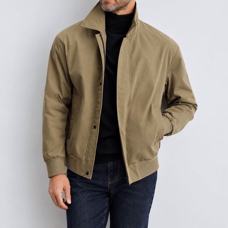 A man wearing a tan utility-style jacket over a black turtleneck sweater and dark blue jeans, photographed against a light gray studio background.