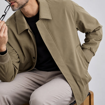 A man wearing an olive green utility jacket over a black t-shirt and light beige trousers, holding a pair of eyeglasses against his face.
