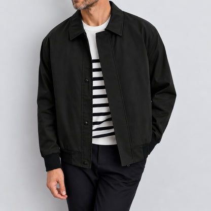 A man wearing a black button-front overshirt jacket over a white and black striped t-shirt and black trousers, photographed against a light gray studio background.