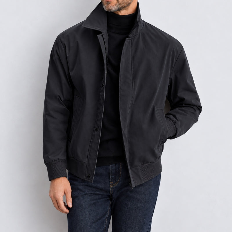 A man wearing a black lightweight bomber-style jacket over a black turtleneck sweater and dark blue jeans, photographed against a plain light gray studio background.