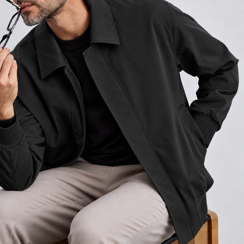 A man wearing a black lightweight jacket over a black t-shirt and beige trousers, seated against a light gray background.