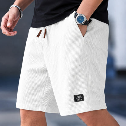 A close-up of a person wearing white textured shorts with a black shirt, a wristwatch, and a bracelet. The shorts feature a subtle grid pattern, brown drawstrings, and a small black brand patch near the hem.