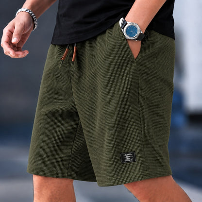 A close-up of a person wearing olive green textured shorts with a black drawstring, a black shirt, a silver watch, and a silver bracelet. The background is a blurred outdoor setting.