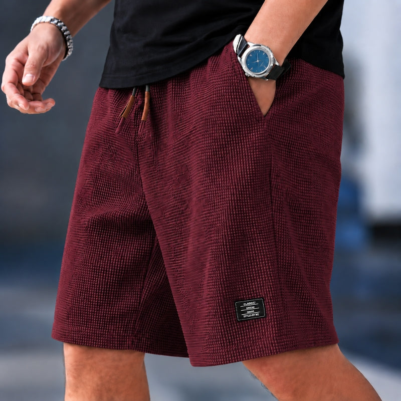 A close-up of a person wearing textured burgundy shorts with a black t-shirt, a silver watch, and a silver bracelet. The shorts feature a subtle grid pattern, a drawstring waist, and a small black brand patch near the hem.