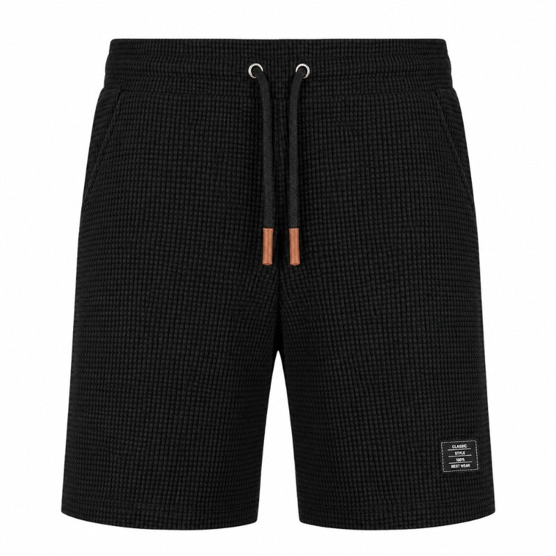 Black textured waffle-knit shorts with a black drawstring featuring brown leather tips and a small white brand label on the lower left leg, shown against a plain white background.