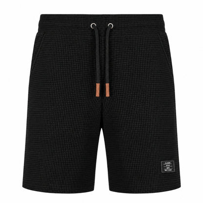 Black textured waffle-knit shorts with a black drawstring featuring brown leather tips and a small white brand label on the lower left leg, shown against a plain white background.