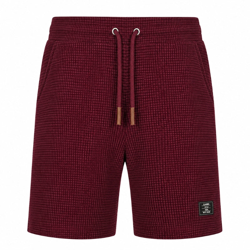 A pair of men's shorts in a textured waffle knit fabric, featuring a deep burgundy color, an elastic waistband with matching drawstrings, and a small black brand patch on the lower left leg.
