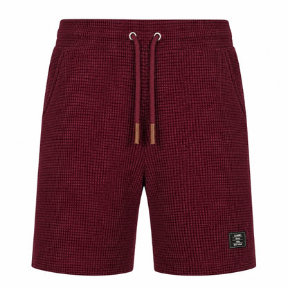 A pair of men's shorts in a textured waffle knit fabric, featuring a deep burgundy color, an elastic waistband with matching drawstrings, and a small black brand patch on the lower left leg.