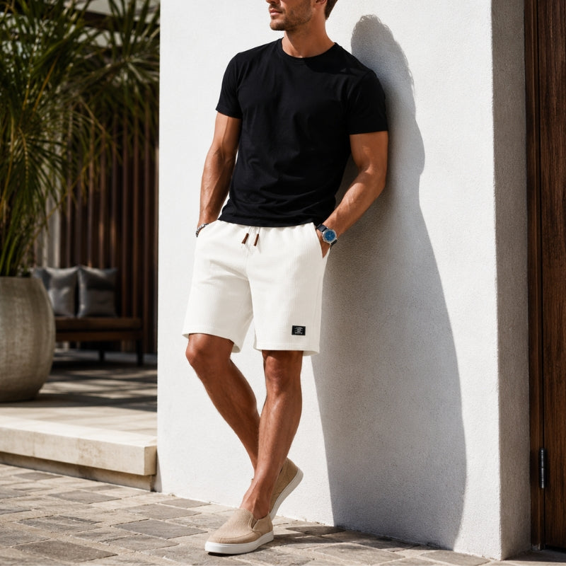 A man wearing a black crewneck t-shirt and off-white drawstring shorts leans against a white stucco wall in a sunny outdoor setting.