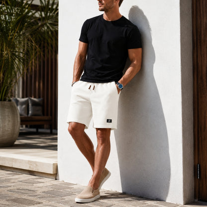 A man wearing a black crewneck t-shirt and off-white drawstring shorts leans against a white stucco wall in a sunny outdoor setting.