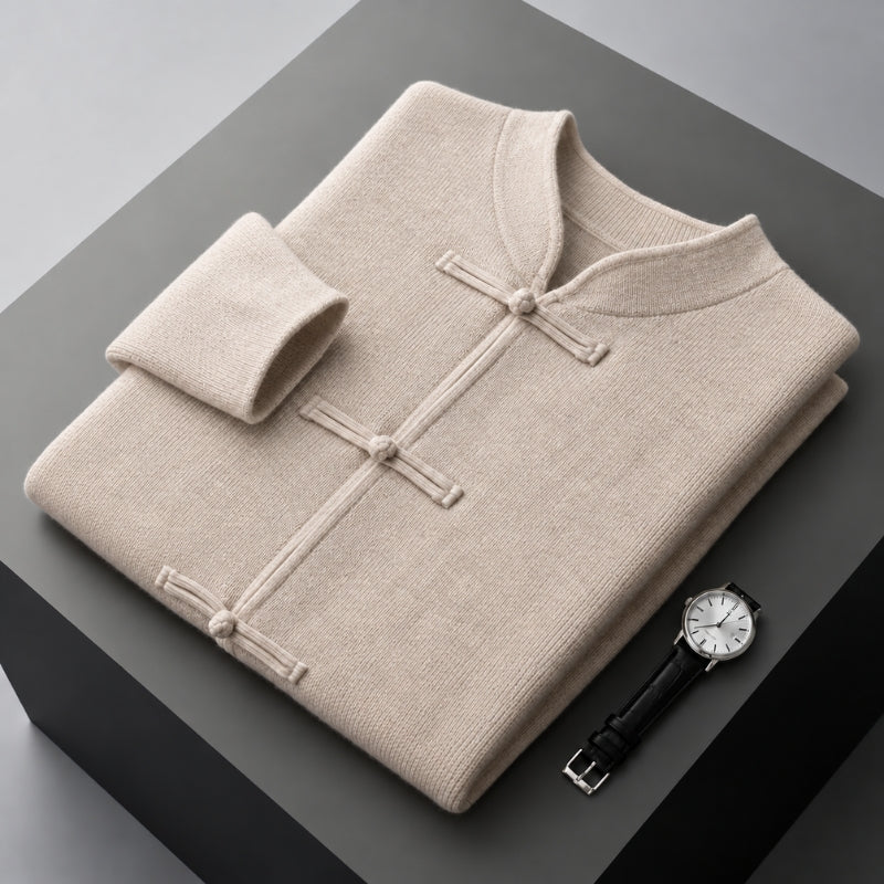 A folded light beige knit sweater with a mandarin collar and button-tab closure, displayed on a dark gray pedestal alongside a wristwatch.