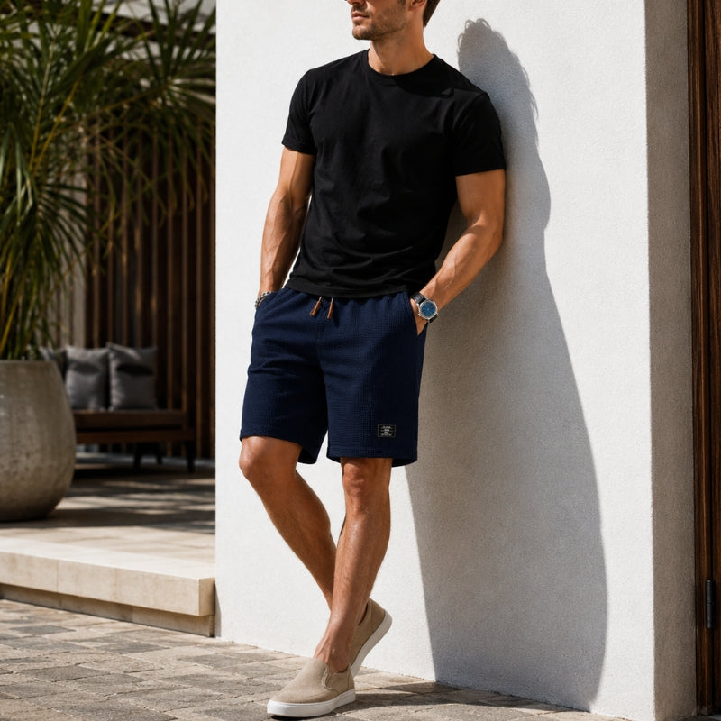 A man wearing a black crewneck t-shirt and navy blue drawstring shorts leans against a white stucco wall in a sunny outdoor setting.
