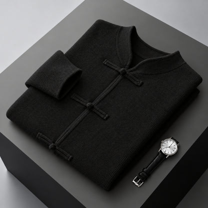 A folded black knit sweater with a mandarin collar and dark button closures, displayed on a dark gray geometric surface next to a wristwatch.