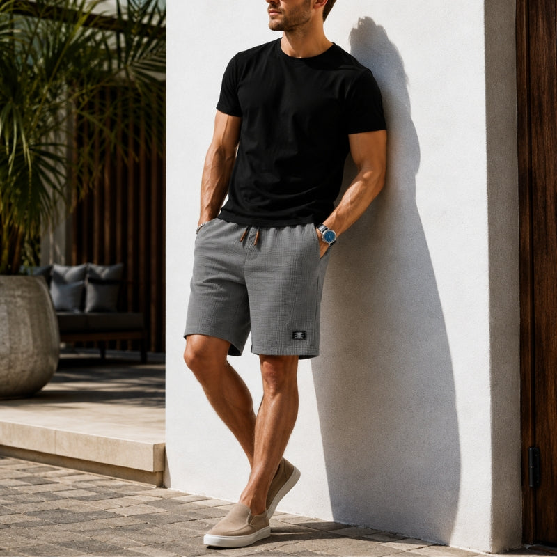 A man wearing gray athletic shorts, a black t-shirt, and beige slip-on shoes leans against a white stucco wall in a sunny outdoor patio setting.