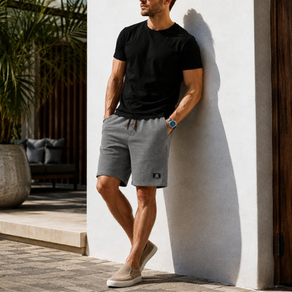 A man wearing gray athletic shorts, a black t-shirt, and beige slip-on shoes leans against a white stucco wall in a sunny outdoor patio setting.