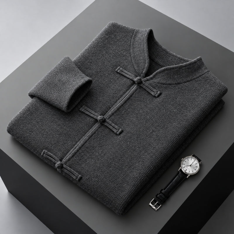 A folded dark gray knit sweater with a mandarin collar and three button closures, displayed on a dark gray pedestal alongside a wristwatch.