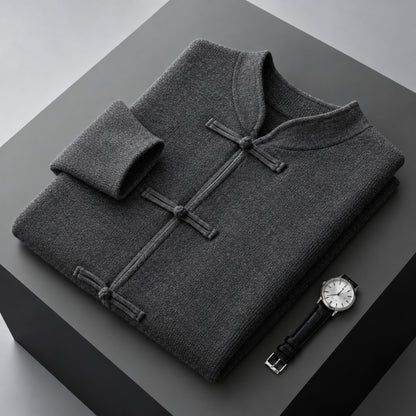 A folded dark gray knit sweater with a mandarin collar and three button closures, displayed on a dark gray pedestal alongside a wristwatch.