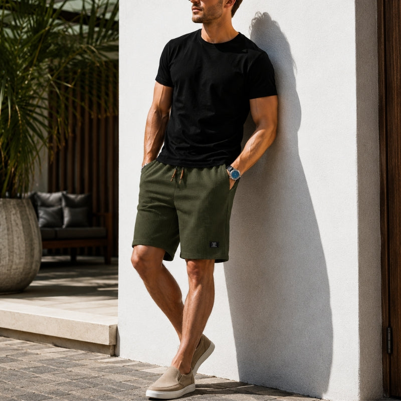 A man wearing olive green drawstring shorts, a black t-shirt, and beige slip-on shoes leans against a white wall in an outdoor patio setting.