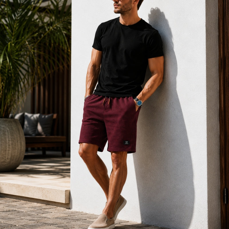 A man wearing a black crewneck t-shirt and burgundy drawstring shorts leans against a white stucco wall in a sunny outdoor setting.