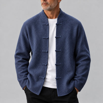 A man wearing a navy blue traditional Chinese-style jacket with fabric toggle closures over a white t-shirt and black pants, photographed against a neutral gray studio background.