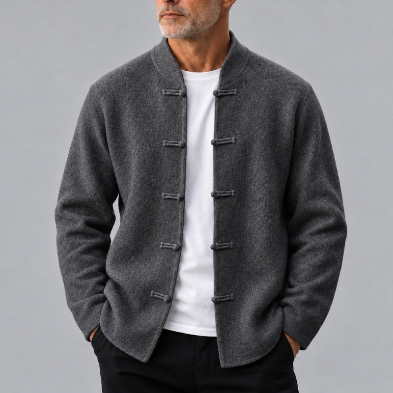 A man wearing a dark gray wool-blend jacket with traditional Chinese-style frog closures over a white t-shirt and black pants, photographed against a solid gray studio background.