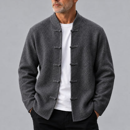 A man wearing a dark gray wool-blend jacket with traditional Chinese-style frog closures over a white t-shirt and black pants, photographed against a solid gray studio background.