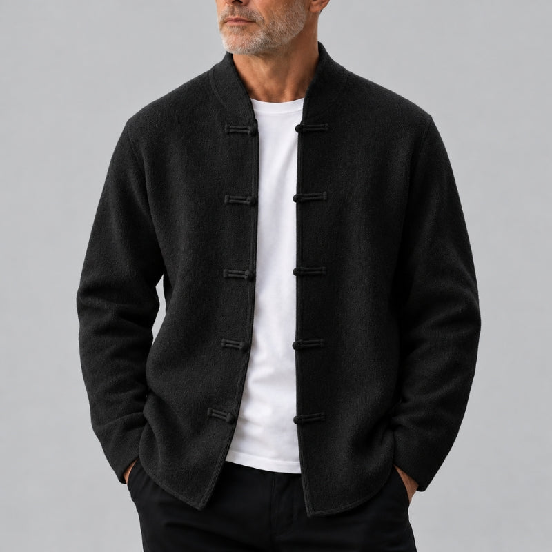A man wearing a black wool-blend jacket with traditional Chinese frog closures, layered over a white t-shirt and black trousers, photographed against a neutral gray background.