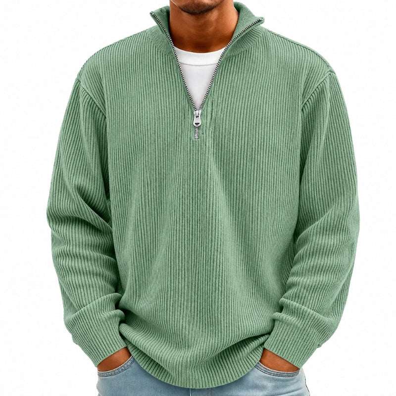 A man wearing a sage green ribbed knit quarter-zip pullover sweater over a white t-shirt and light blue jeans, shown from the chin down against a plain white background.