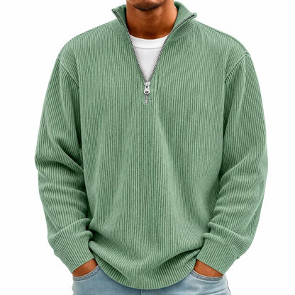 A man wearing a sage green ribbed knit quarter-zip pullover sweater over a white t-shirt and light blue jeans, shown from the chin down against a plain white background.
