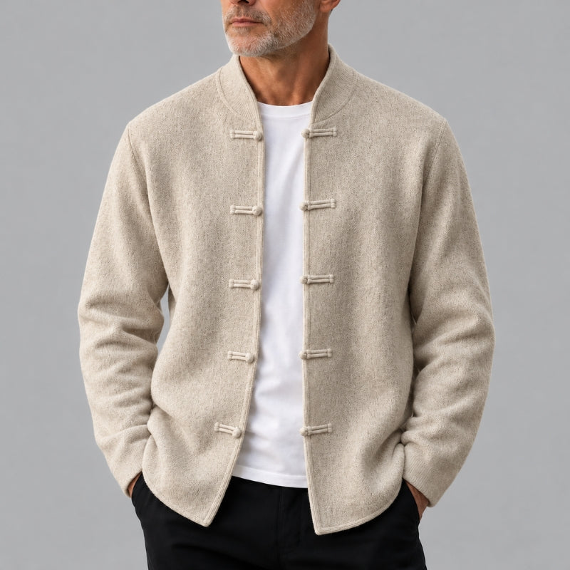 A man wearing a light beige textured Chinese-style mandarin collar jacket with toggle closures over a white t-shirt and black pants, photographed against a solid gray studio background.