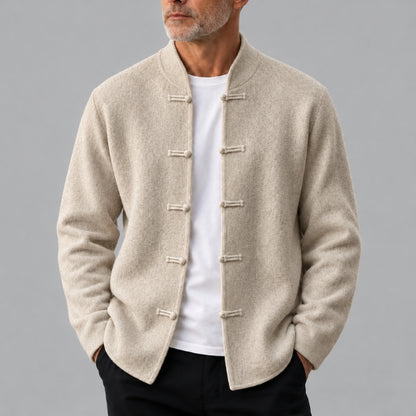 A man wearing a light beige textured Chinese-style mandarin collar jacket with toggle closures over a white t-shirt and black pants, photographed against a solid gray studio background.