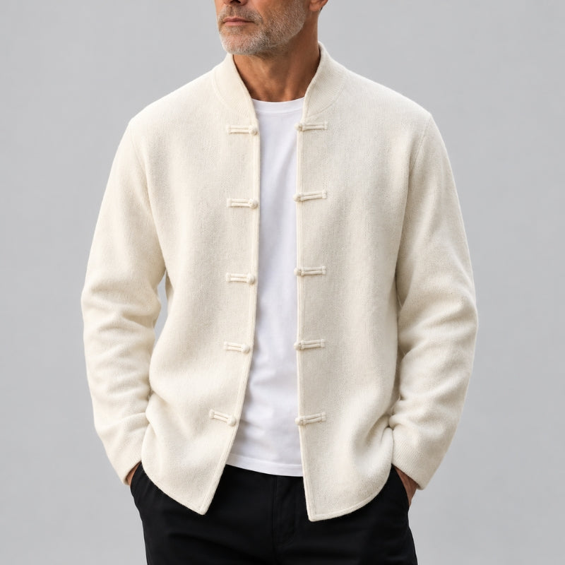 A man wearing a cream-colored Chinese-style jacket with toggle closures over a white t-shirt and black pants, photographed against a neutral gray background.