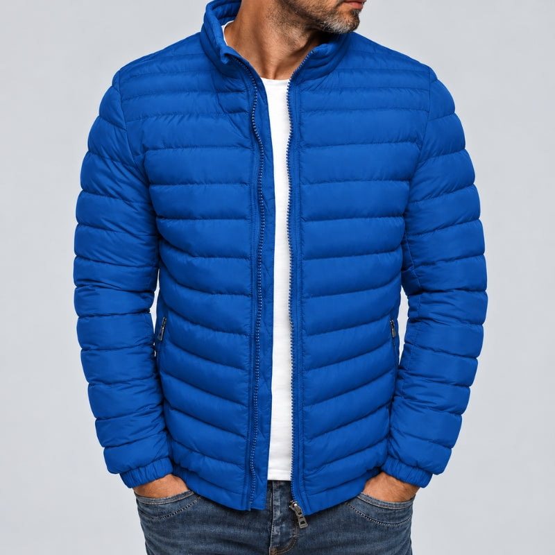 A man wearing a bright royal blue quilted puffer jacket over a white t-shirt and dark blue jeans, shown from the chin down against a solid gray background.