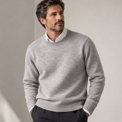 A man wearing a light gray crewneck sweater over a white collared shirt, standing against a neutral beige background.