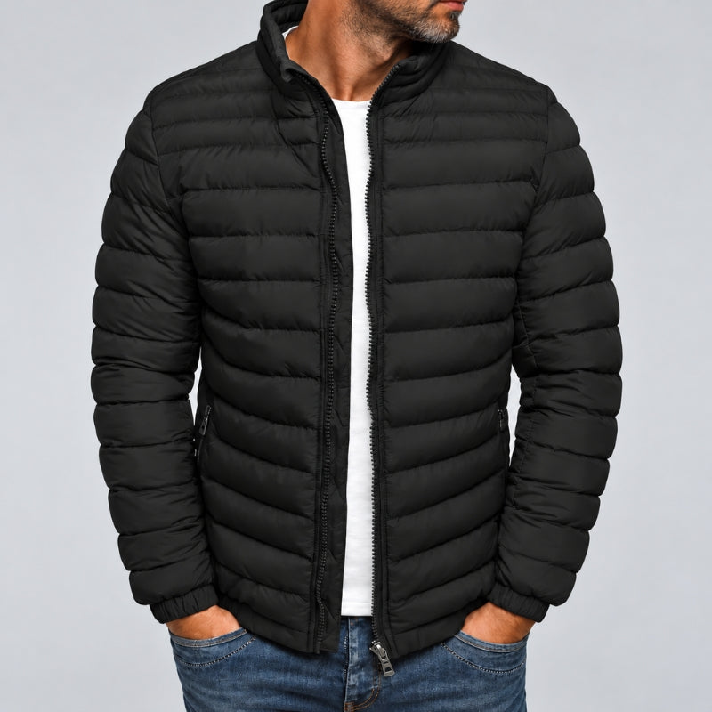 A man wearing a black puffer jacket with horizontal quilting, a stand-up collar, and a front zipper, layered over a white shirt and blue jeans, against a plain light gray background.