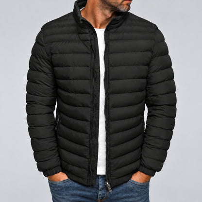A man wearing a black puffer jacket with horizontal quilting, a stand-up collar, and a front zipper, layered over a white shirt and blue jeans, against a plain light gray background.