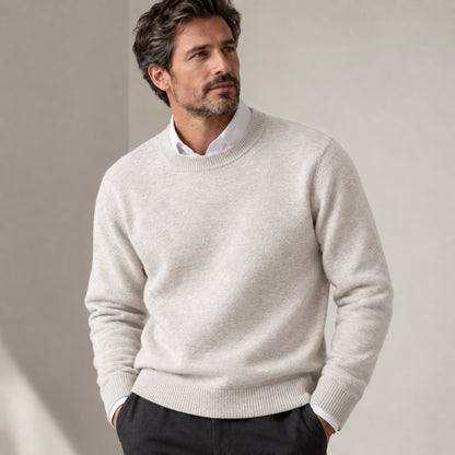A man wearing a light beige crew-neck knit sweater over a white collared shirt and dark trousers, standing against a neutral beige wall.