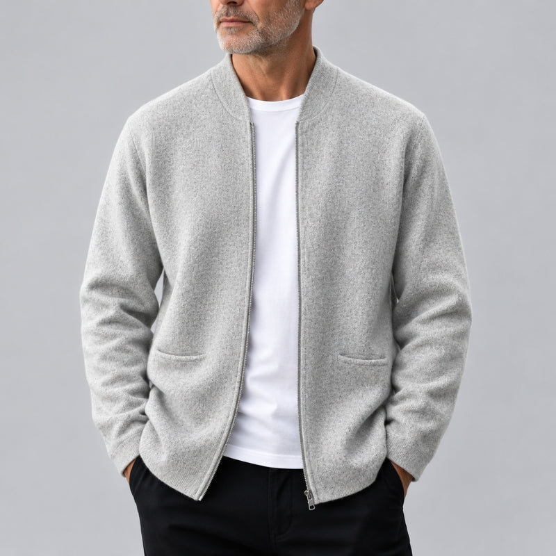 A man wearing a light gray zip-up knit cardigan over a white t-shirt and black pants, shown from the chin down against a solid gray studio background.