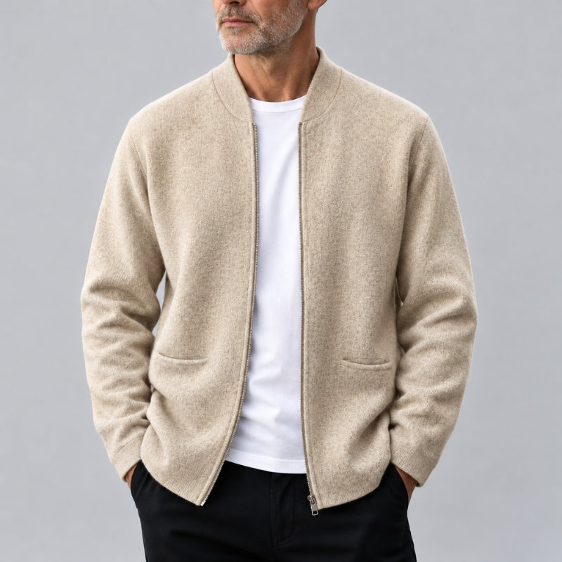 A man wearing a light beige zip-up knit cardigan over a white t-shirt and black pants, photographed against a neutral gray studio background.