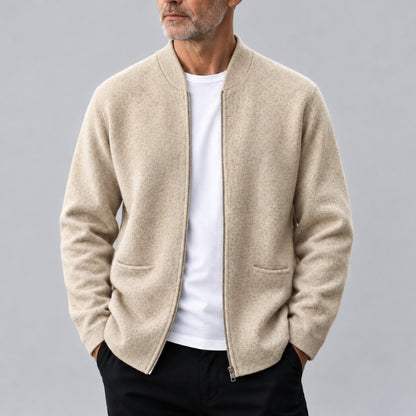 A man wearing a light beige zip-up knit cardigan over a white t-shirt and black pants, photographed against a neutral gray studio background.