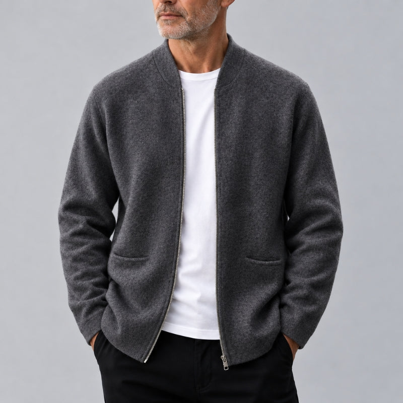 A man wearing a dark gray zip-up knit jacket over a white t-shirt and black pants, photographed against a neutral gray studio background.