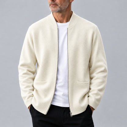 A man wearing a cream-colored zip-up knit jacket over a white t-shirt and black pants, photographed against a neutral gray studio background.