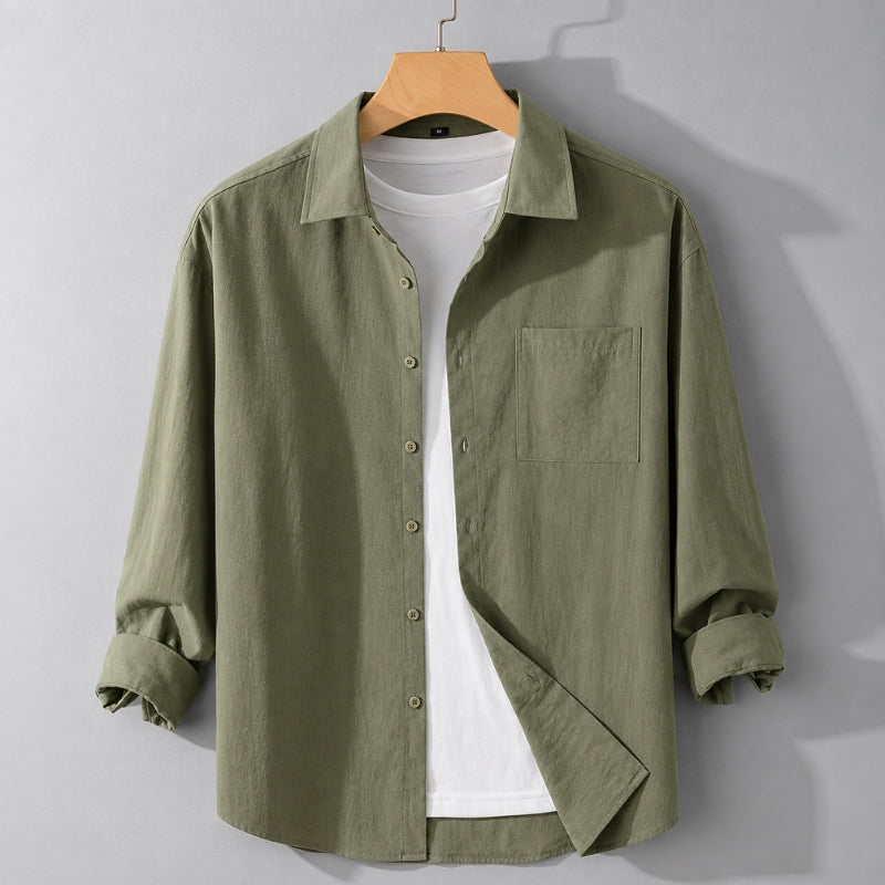 An olive green long-sleeve linen button-up shirt with a chest pocket, styled over a white t-shirt on a wooden hanger against a gray wall.
