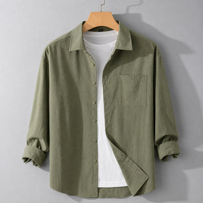 An olive green long-sleeve linen button-up shirt with a chest pocket, styled over a white t-shirt on a wooden hanger against a gray wall.