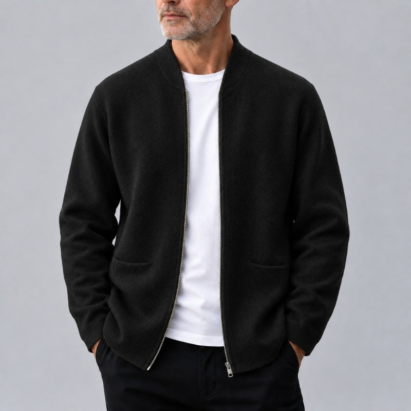 A man wearing a black zip-up bomber jacket over a white t-shirt and black pants, standing against a solid gray studio background.