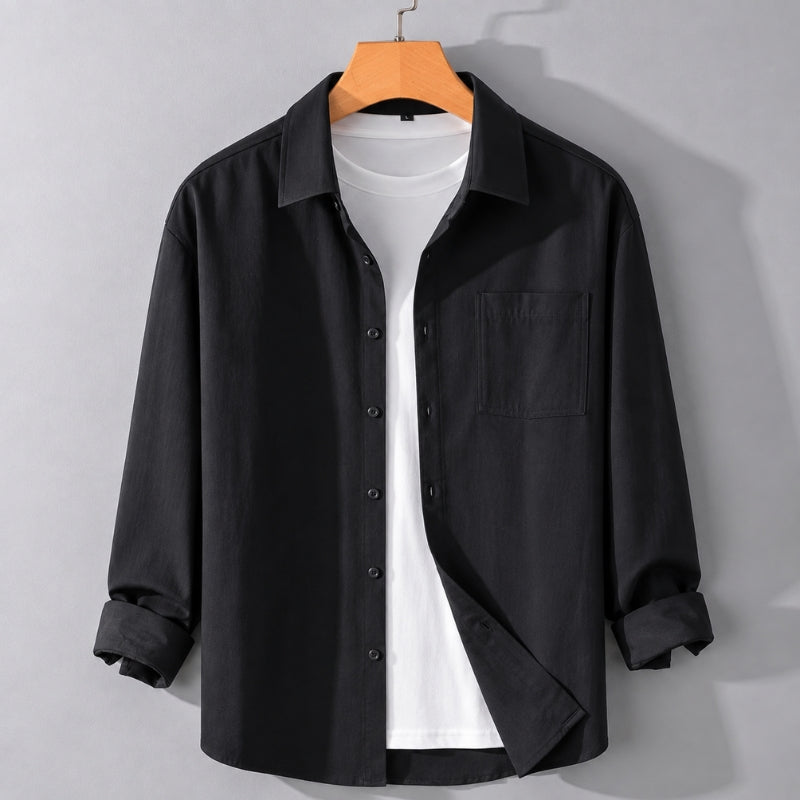 A black long-sleeve button-up shirt with a chest pocket, styled open over a white t-shirt and hanging on a wooden hanger against a light gray wall.