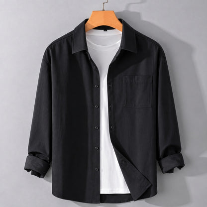 A black long-sleeve button-up shirt with a chest pocket, styled open over a white t-shirt and hanging on a wooden hanger against a light gray wall.