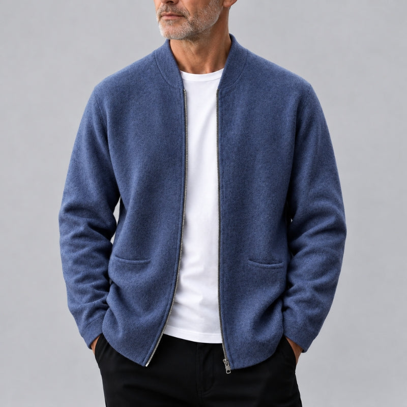 A man wearing a blue zip-up knit jacket over a white t-shirt and black pants, photographed against a plain gray studio background.