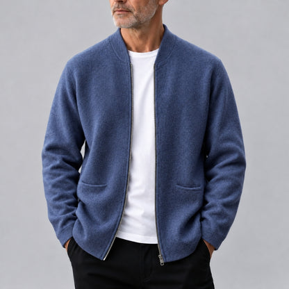 A man wearing a blue zip-up knit jacket over a white t-shirt and black pants, photographed against a plain gray studio background.