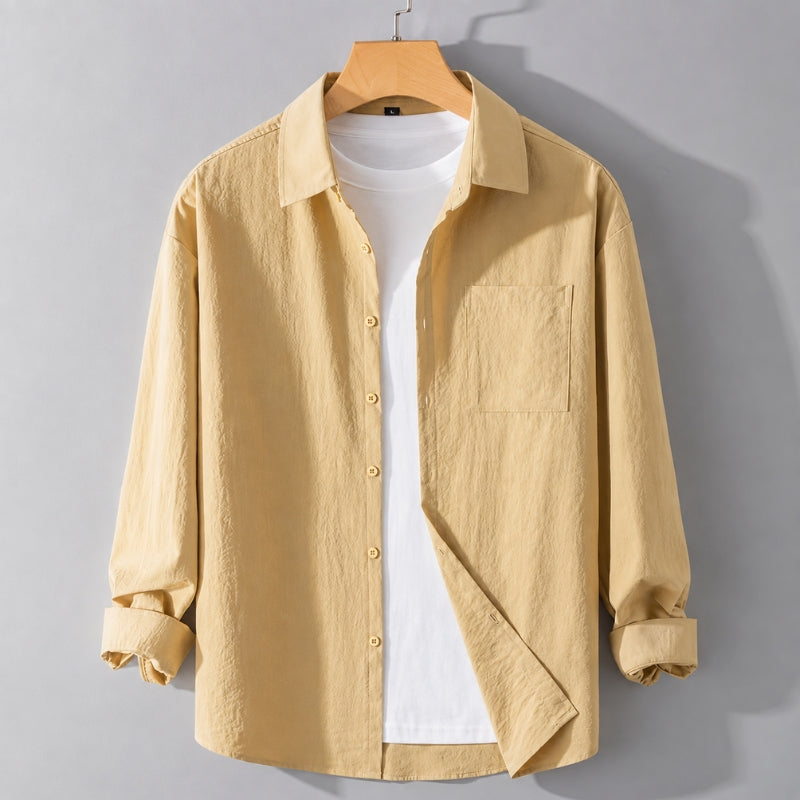 A light yellow long-sleeve button-up shirt with a chest pocket, styled over a white t-shirt on a wooden hanger against a gray background.